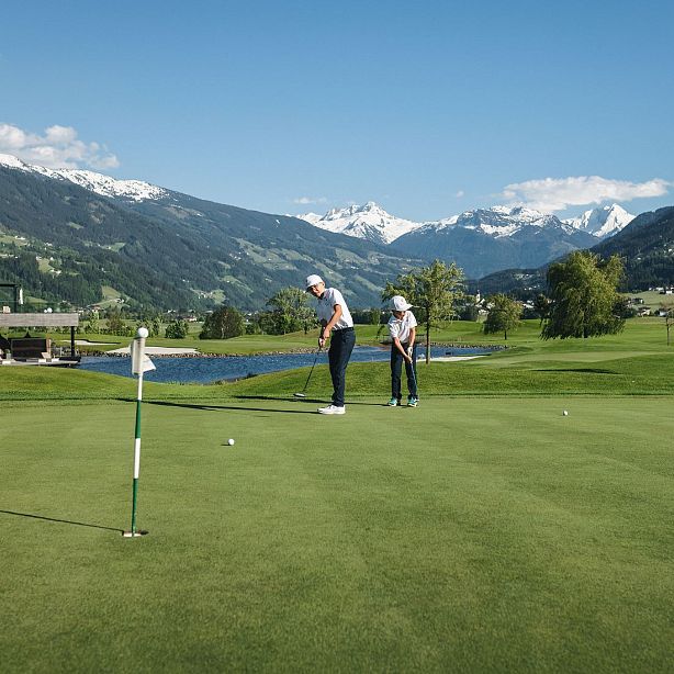 Zwei Personen spielen Golf auf einem großen, grünen Golfplatz mit Bergkulisse. Der Himmel ist blau und sonnig, und im Hintergrund sind schneebedeckte Gipfel zu sehen.