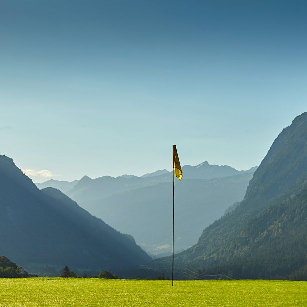 A golf course with a yellow flag in a lush valley, flanked by tall mountains under a clear blue sky.