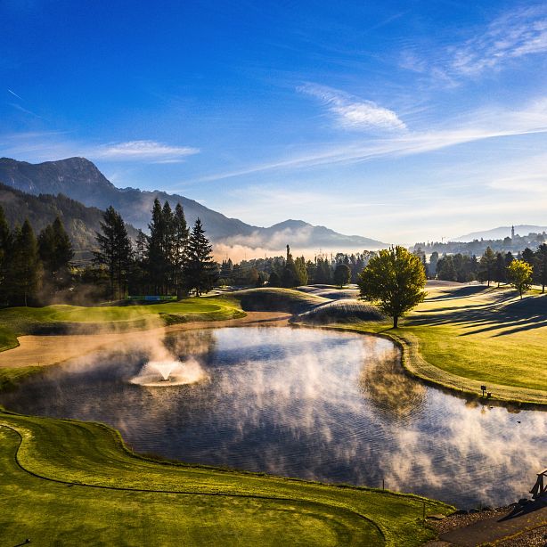 Scenic view of a golf course at dawn with a misty pond, lush greenery, and mountains under a clear sky.