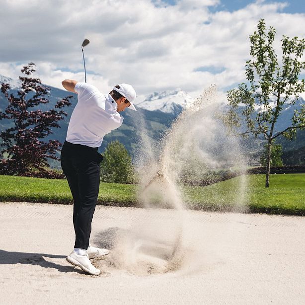 Ein Golfer schlägt mit seinem Schläger aus einem Sandbunker. Sand wirbelt in die Luft. Im Hintergrund sind Berge und blauer Himmel zu sehen.