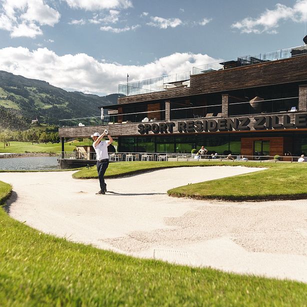 Ein Golfer schlägt einen Ball auf einem gepflegten Platz. Im Hintergrund steht ein modernes Gebäude mit der Aufschrift "Sport Residenz Zillertal".