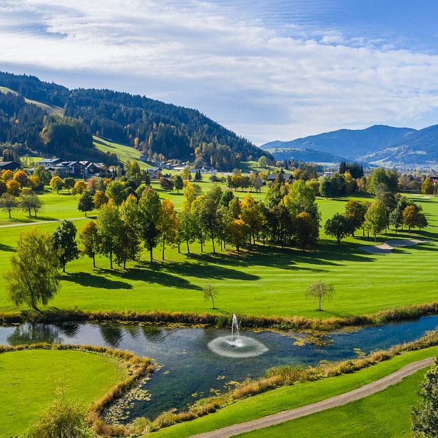 Lush green golf course with trees and a small pond with a fountain, surrounded by mountains under a bright blue sky.