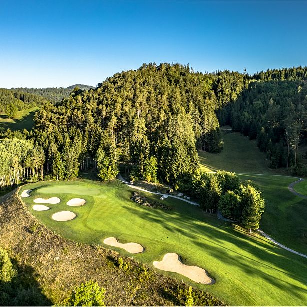 Aerial view of a golf course with green fairways and sand bunkers, surrounded by dense forest and rolling hills under a clear blue sky.