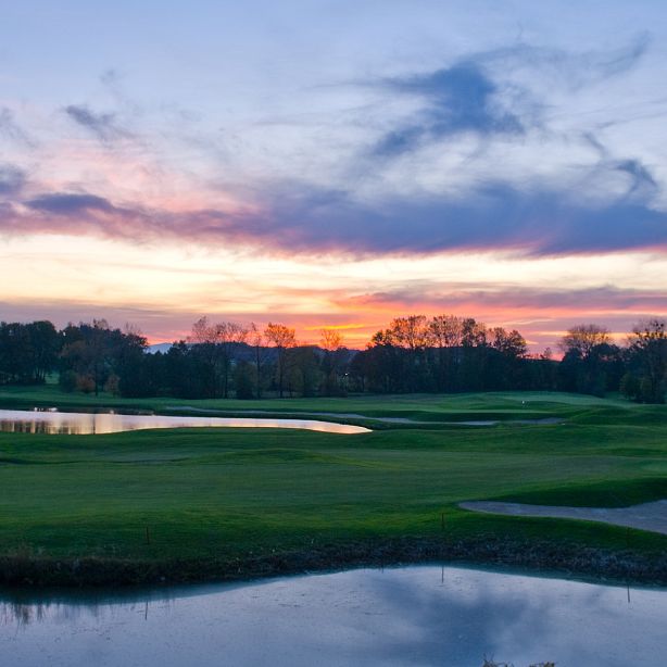 Ein malerischer Sonnenuntergang über einem Golfplatz. Der Himmel ist in lebendigen Rosa- und Blautönen gefärbt, während sich Wasserflächen im Vordergrund spiegeln.