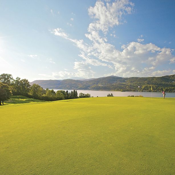 Golf course overlooking a lake with mountains in the background, bright sun and blue sky, surrounded by trees and lush vegetation.