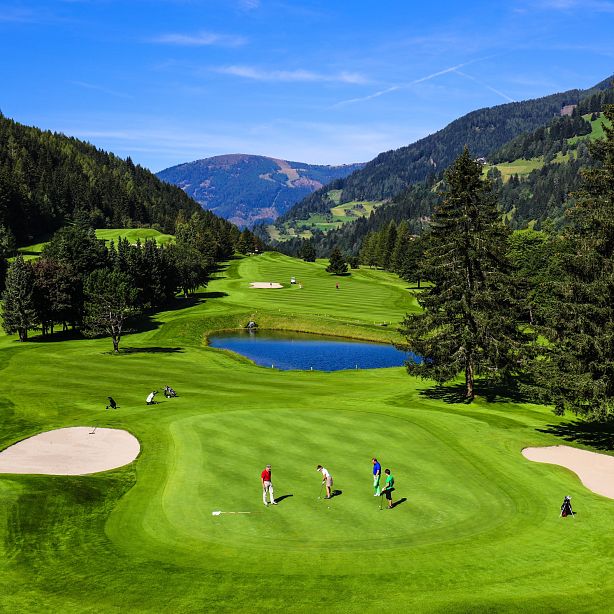 Golf course in a valley with lush greens, sand bunkers, a small pond, and golfers playing. Surrounded by mountains and tall trees under a clear blue sky.