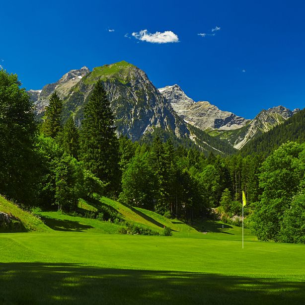 Grüne Golfbahn umgeben von dichtem Wald, dahinter eindrucksvolle Bergkulisse unter strahlend blauem Himmel. Eine Fahne markiert das Loch auf dem Grün.