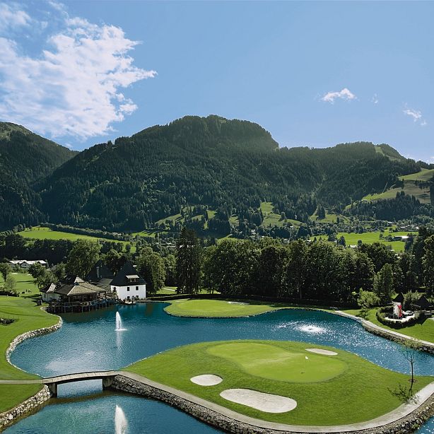 Panorama eines Golfplatzes inmitten einer bergigen Landschaft mit einem Teich im Vordergrund, umgeben von grünen Wiesen und bewaldeten Hügeln unter blauem Himmel.