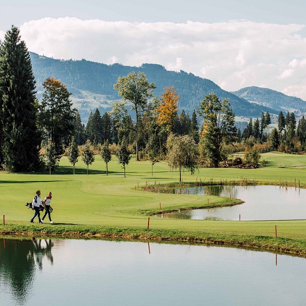 Zwei Menschen spazieren mit Golfbags über einen gepflegten Golfplatz mit mehreren Teichen im Vordergrund und bewaldeten Hügeln im Hintergrund.