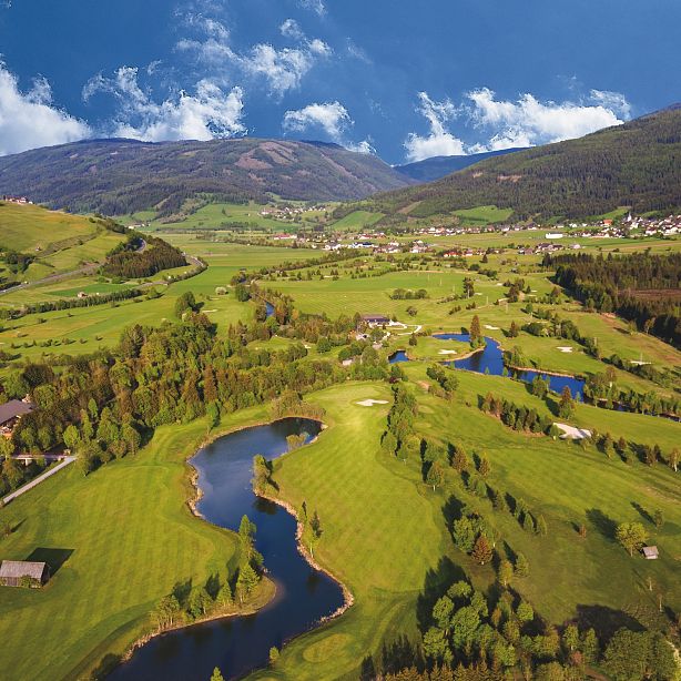 Luftaufnahme einer weitläufigen, grünen Landschaft mit Fluss, Wäldern und Bergen im Hintergrund. Ein kleiner Ort ist in der Ferne sichtbar.