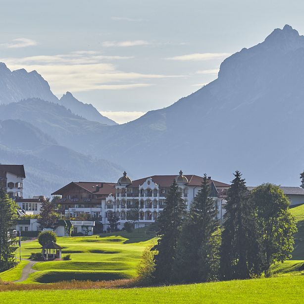 Ein malerisches Landschaftsbild zeigt ein Hotel in den Alpen, umgeben von grünen Wiesen und hohen Bergen im Hintergrund, unter einem bewölkten Himmel.