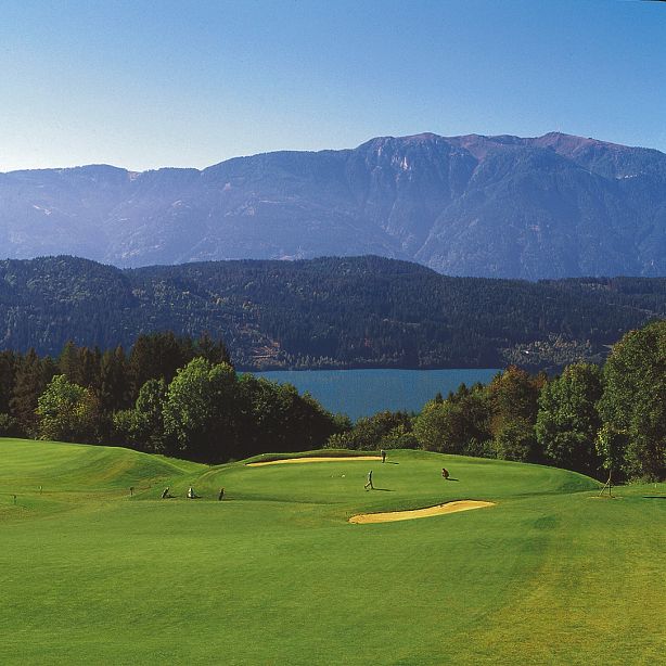 Malere Kulisse eines Golfplatzes mit grünen Fairways und einem Sandbunker. Im Hintergrund sind ein blauer See und hohe, bewaldete Berge zu sehen.