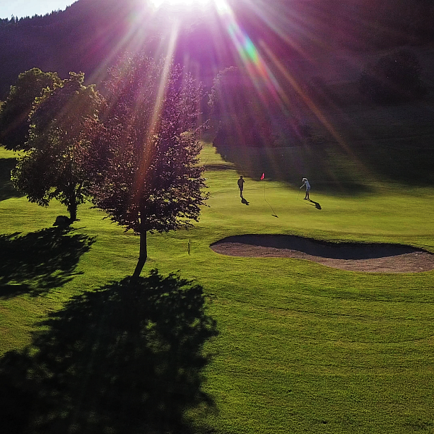 Golflandschaft bei Sonnenuntergang mit einer grünen Wiese, Bäumen und einem Sandbunker, beleuchtet von der warmen Abendsonne, die durch die Bäume scheint.