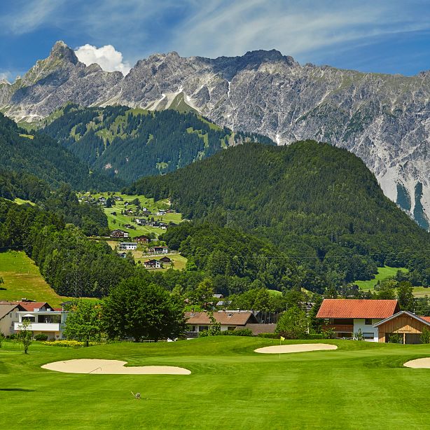 A picturesque golf course with green fairways, sand bunkers, and quaint houses is set against majestic alpine mountains under a blue sky.