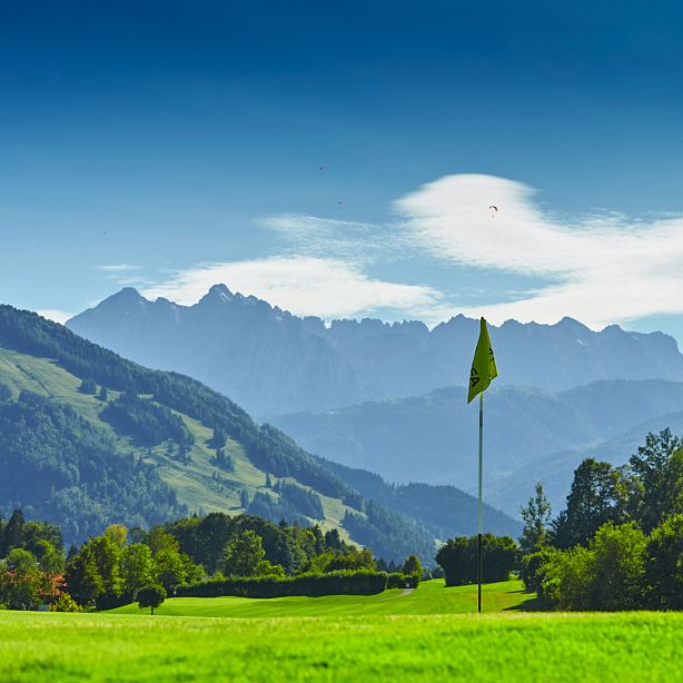 Grüne Wiesen mit einem Golfplatz, dahinter majestätische Berge unter blauem Himmel. Vereinzelte Wolken verleihen dem Bild eine friedliche Atmosphäre.