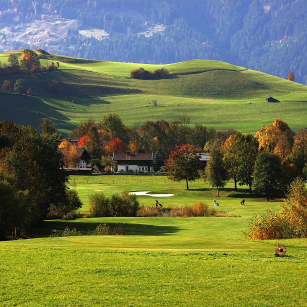 Lush golf course in a hilly landscape with autumn-colored trees and distant mountains. Players are visible, enjoying a serene game under clear skies.