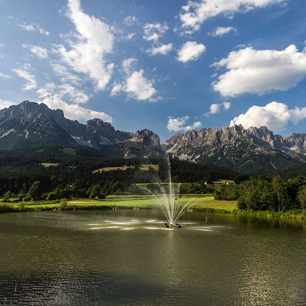 Berglandschaft mit einem See im Vordergrund, einer sprudelnden Fontäne und majestätischen Bergen im Hintergrund unter wolkigem Himmel.