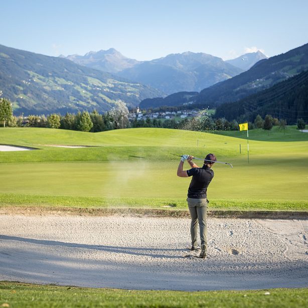 Golfer schlägt im Bunker eines gepflegten Golfplatzes ab, umgeben von hügeligen, bewaldeten Bergen und Wolken am blauen Himmel. Fahne im Hintergrund sichtbar.