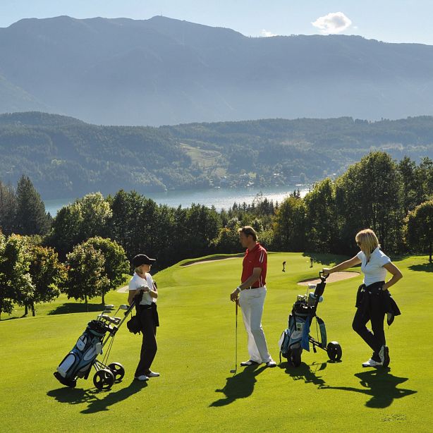 Drei Golfspieler stehen auf einem grünen Platz unter blauem Himmel, umgeben von Bergen und Bäumen. Sie diskutieren, während sie auf ihre Schläger gestützt sind.