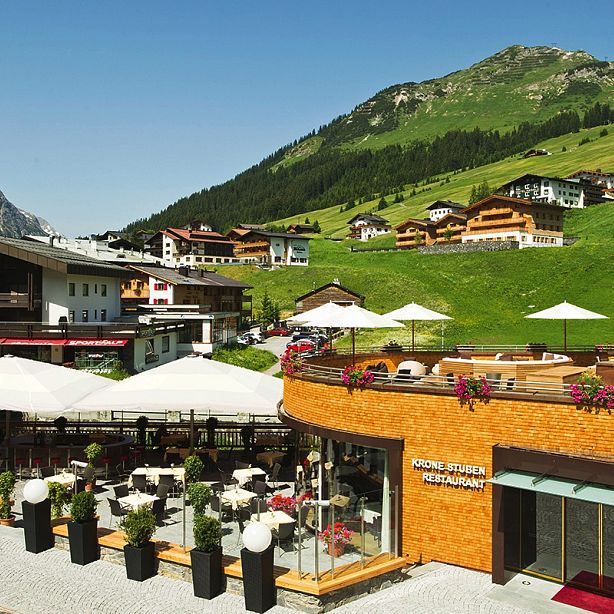 Ein luxuriöses Hotel mit Terrasse in alpiner Umgebung, umgeben von grünen Wiesen und Bergen unter einem klaren blauen Himmel, ist zu sehen.