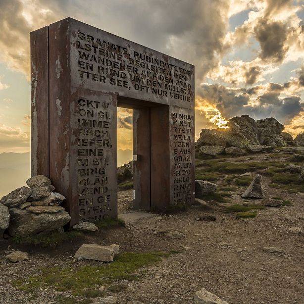 A stone portal with engraved text stands on a rocky mountain peak at sunset, surrounded by clouds and rugged terrain.