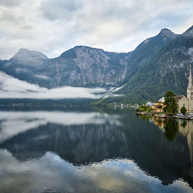 A tranquil lake reflects mountains and a village with a church; misty clouds hover over the picturesque landscape, creating a serene atmosphere.