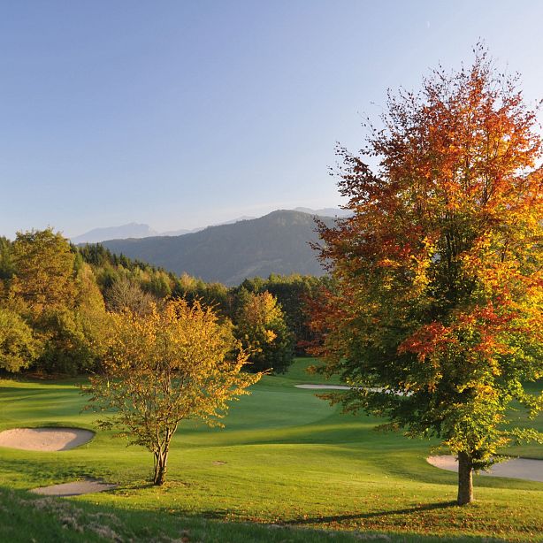 Bunte Herbstlandschaft auf einem Golfplatz; leuchtend gefärbte Bäume umgeben die gepflegten grünen Fairways und Sandbunker, darunter blauer Himmel.