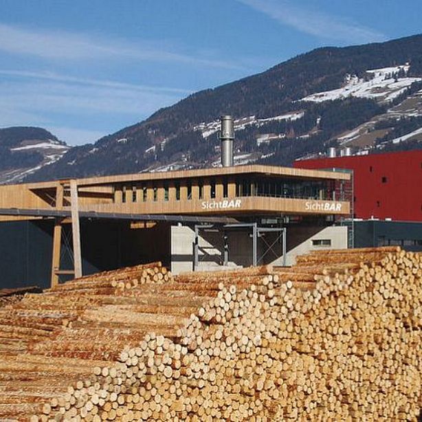 Großes Holzlager vor einem modernen Gebäude aus Holz und Glas, mit der Aufschrift "SichtBAR". Im Hintergrund sind schneebedeckte Berge zu sehen.