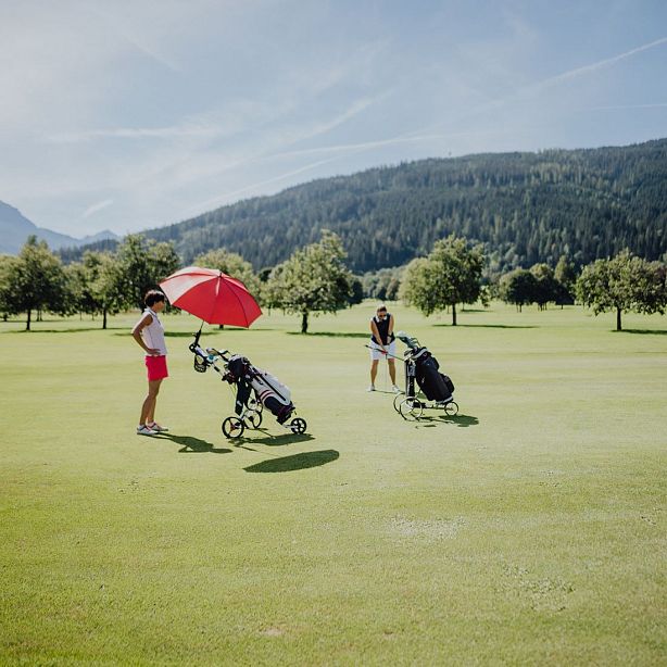 Two golfers on a green course; one with a red umbrella. Trees and mountains are in the sunny background.