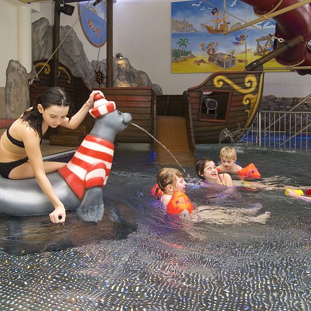 Children and adults enjoy swimming in an indoor pool with a seal float and a pirate ship background.
