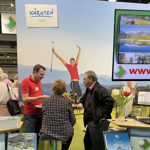 A man in a red shirt speaks with two people at a tourism event booth. Brochures and promotional materials are displayed around them.