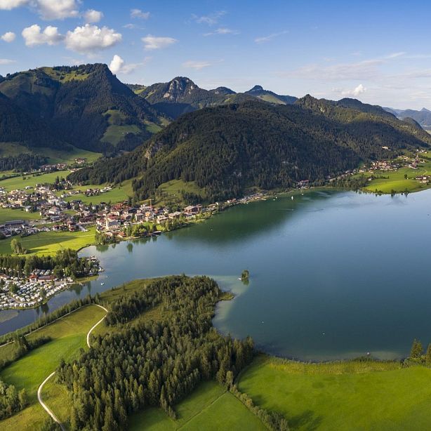 Aerial view of a tranquil Alpine lake surrounded by green hills, a small town, and mountains under a clear sky.