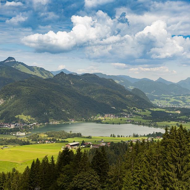 Scenic view of green hills, a lake, and mountains under a partly cloudy sky in an alpine region.