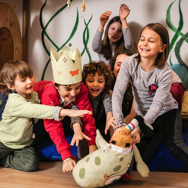 Eine Gruppe fröhlicher Kinder spielt zusammen in einem Raum mit Holzmöbeln und Wandmalereien. Ein Kind trägt eine Krone aus Papier.