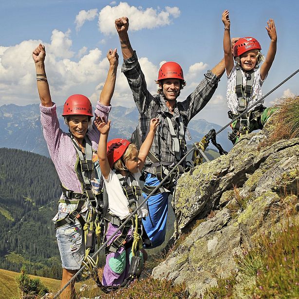 Eine Gruppe von vier Personen, alle in Kletterausrüstung und Helmen, steht fröhlich auf einem Bergpfad mit erhobenen Armen. Im Hintergrund sind Berge zu sehen.