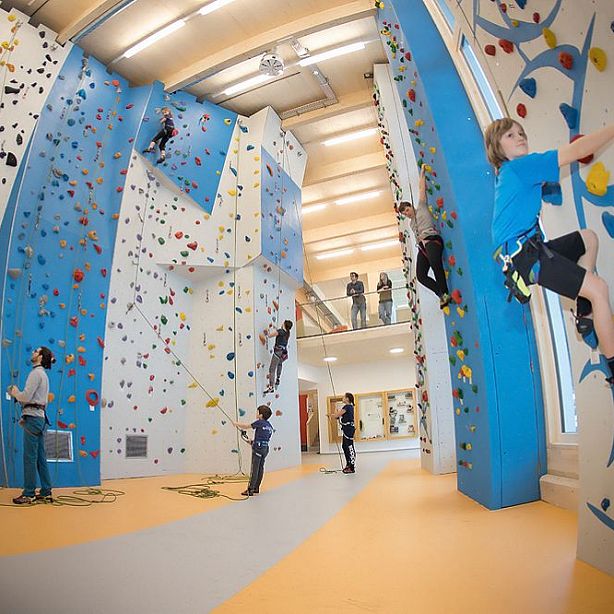 Mehrere Menschen klettern an einer großen, modernen Kletterwand in einer Sporthalle. Kinder und Erwachsene tragen Kletterausrüstung und sichern sich gegenseitig.