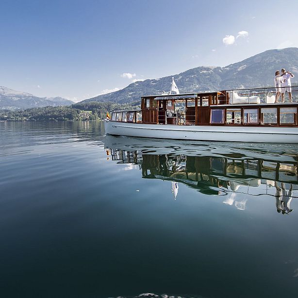 A boat on a calm lake with mountains in the background, under a clear sky. The water reflects the boat and surroundings, creating a serene scene.