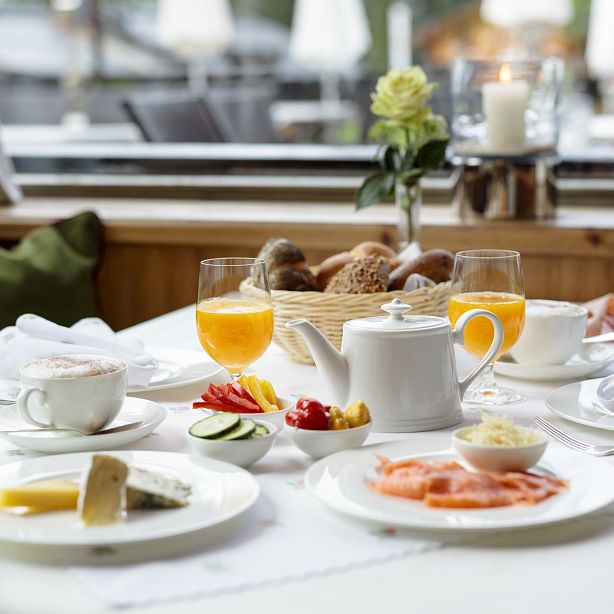 Ein gedeckter Frühstückstisch mit Orangensaft, Tee, Brot, Käse, Lachs und Gemüse. Im Hintergrund eine Kerze und eine Blume zur Dekoration.