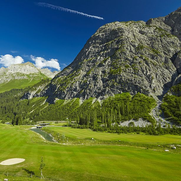 Golfplatz in alpiner Landschaft mit saftig grünem Rasen, umgeben von beeindruckenden Bergen und dichten Wäldern unter einem klaren blauen Himmel.