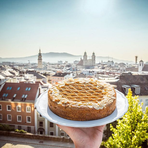 A cityscape with historic buildings under a clear sky, highlighted by a cake topped with almonds in the foreground on a white platter.