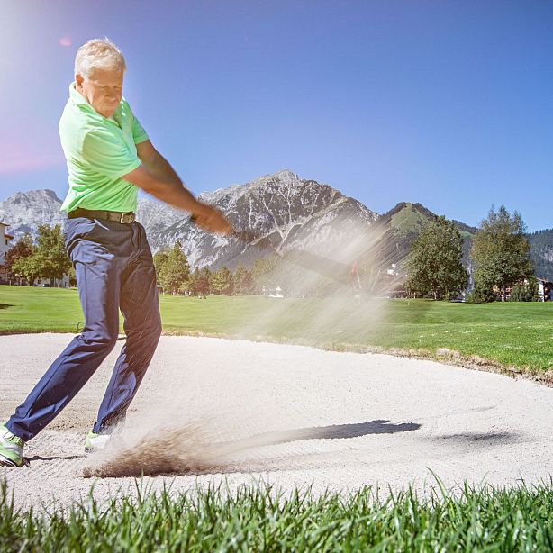 A golfer in a green shirt and blue pants hits a sand shot in a golf course with mountains and trees under a clear blue sky.