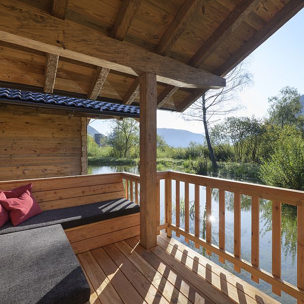Wooden cabin veranda with seating overlooks calm lake and distant hills, surrounded by lush greenery under a clear blue sky.