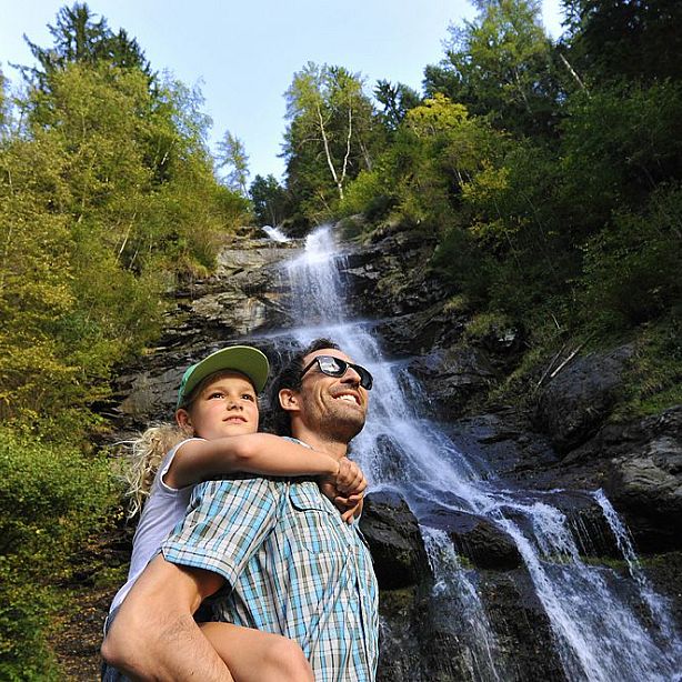 Ein Mann mit Sonnenbrille trägt ein Kind auf dem Rücken. Beide lächeln. Im Hintergrund ein malerischer Wasserfall und viel Grün durch Bäume und Pflanzen.