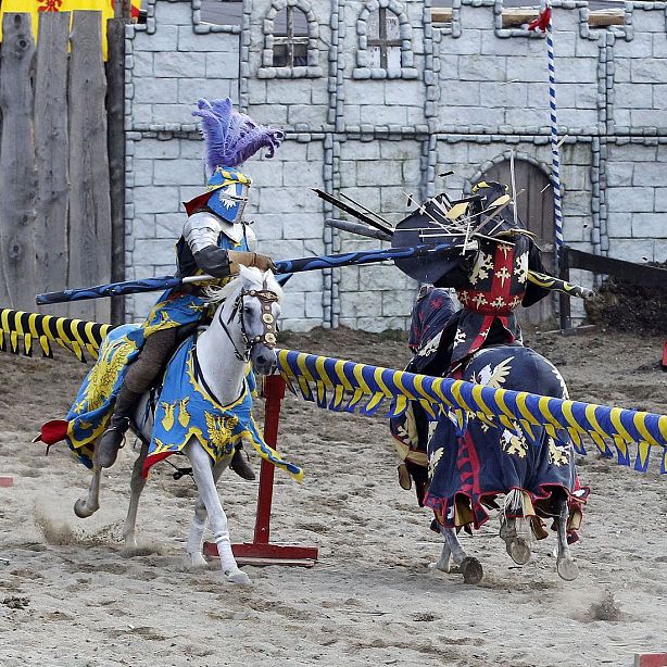 Two knights in colorful armor joust on horseback in a sandy arena with a stone castle wall behind them. They are mid-action, striking each other with lances.