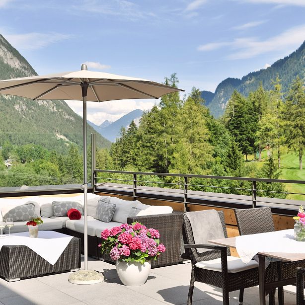 Terrasse mit gemütlichen Sitzmöbeln, Blumen und Sonnenschirmen, umgeben von grünen Wiesen und majestätischen Bergen unter einem klaren blauen Himmel.