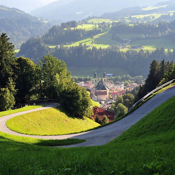 A beautiful mountain village nestled in lush green hills, with a winding road and a clear view of the distant valley and a church in the center.