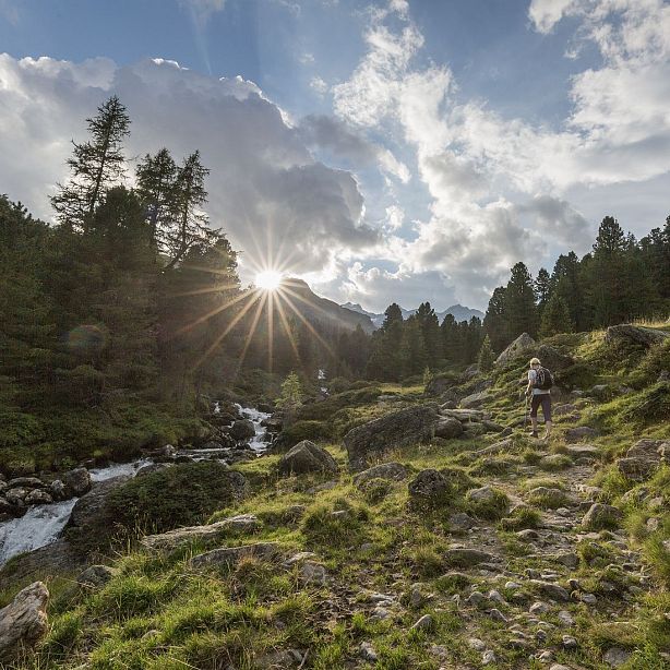 A hiker on a rocky path in a mountain landscape at sunrise, with trees and a stream under a sunlit sky filled with clouds.