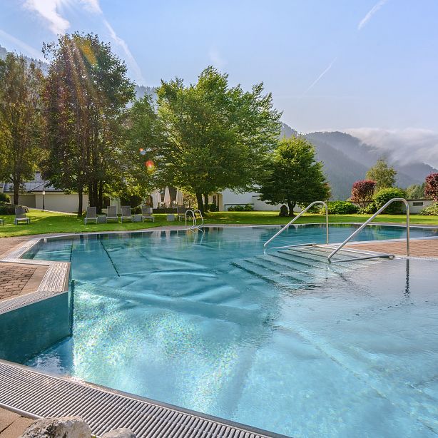 Outdoor pool with clear blue water, surrounded by lush greenery and mountains under a clear sky, creating a serene and tranquil atmosphere.