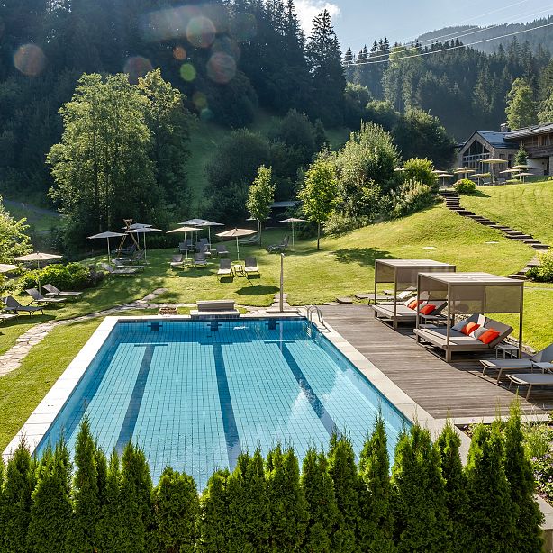 Ein idyllisches Landschaftsbild zeigt einen rechteckigen Pool, umgeben von Liegestühlen und Sonnenschirmen, eingebettet in einer grünen, waldreichen Umgebung.