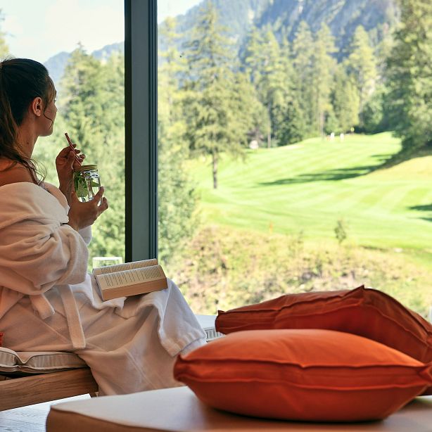 Woman in a robe sits by a window, holding a drink and a book, gazing at a lush green landscape with trees and mountains in the distance.